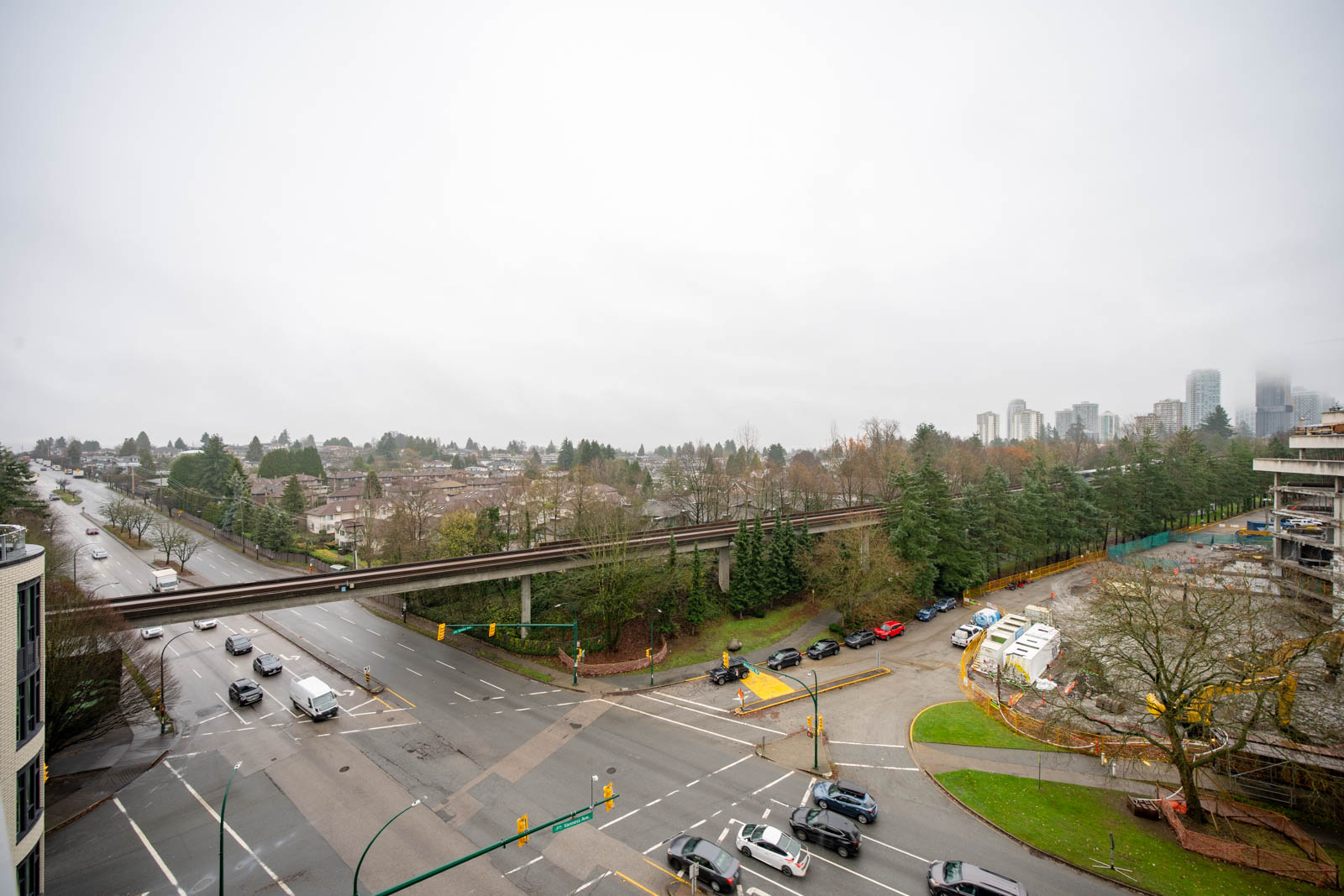 A wide intersection with cars, a light rail track running overhead, and a parking lot on the right, under an overcast sky in an urban area.