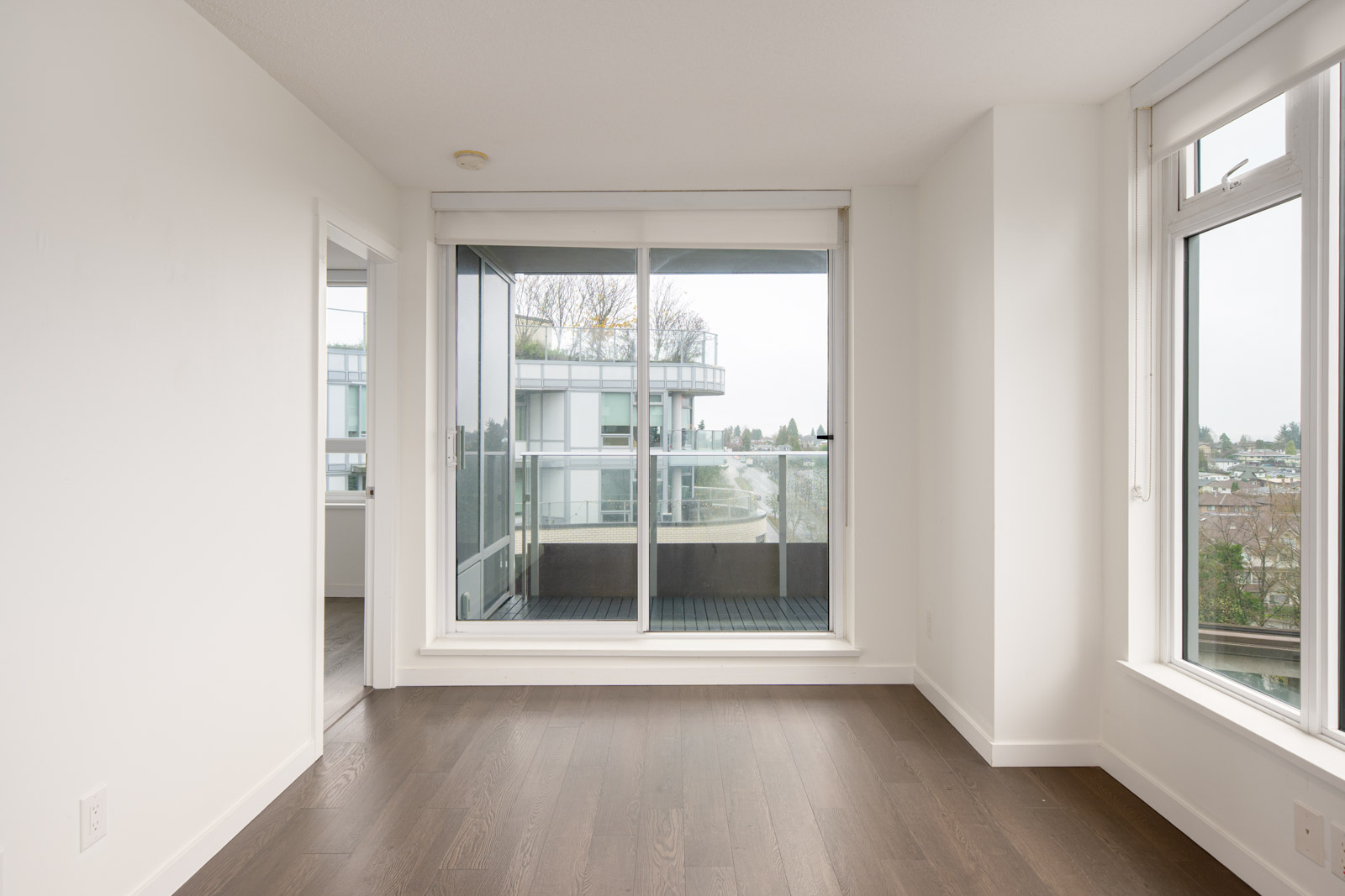 Empty room with dark wood floor, white walls, and large windows with a sliding glass door leading to a balcony overlooking nearby buildings and trees.