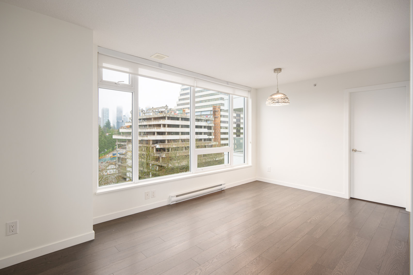 Unfurnished room with large windows, hardwood floor, white walls, a ceiling light fixture, and a view of modern buildings outside.