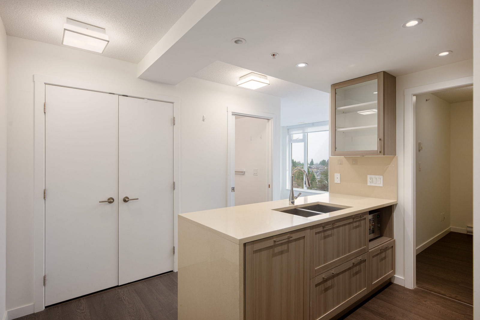 Modern apartment corner with a small kitchen area featuring a sink, wood cabinetry, and cream countertop; two white double doors and a window are visible in the background.