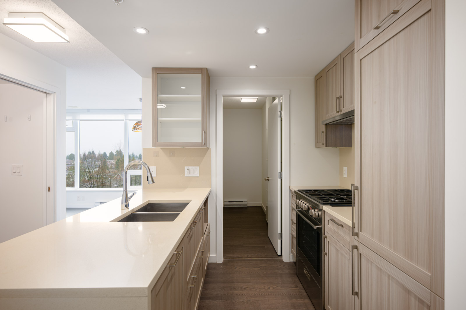 Modern kitchen with light wood cabinets, white countertops, double sink, built-in oven, and gas stove; hallway and bright windowed room visible in the background.