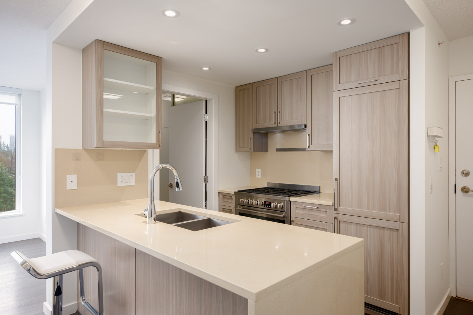 Modern kitchen with light wood cabinets, built-in appliances, a beige countertop island with a sink, bar stool, and recessed ceiling lights.