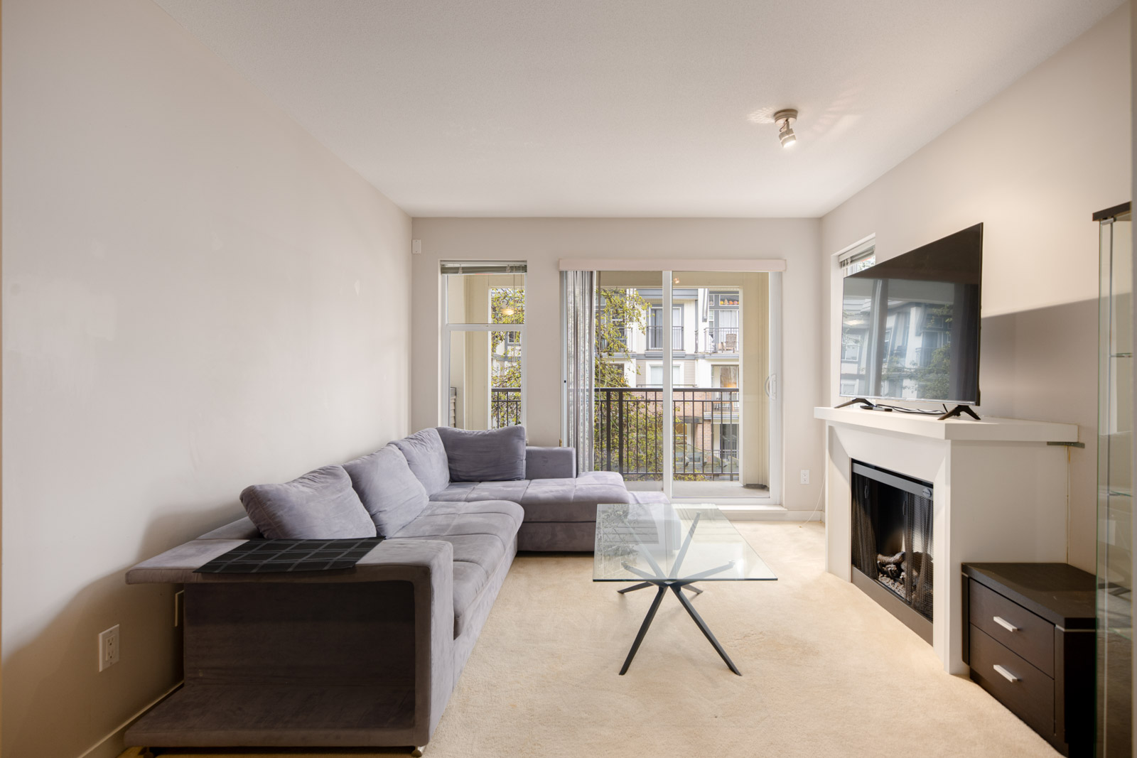 Modern living room with a gray sectional sofa, glass coffee table, mounted TV above a fireplace, and windows leading to a small balcony.