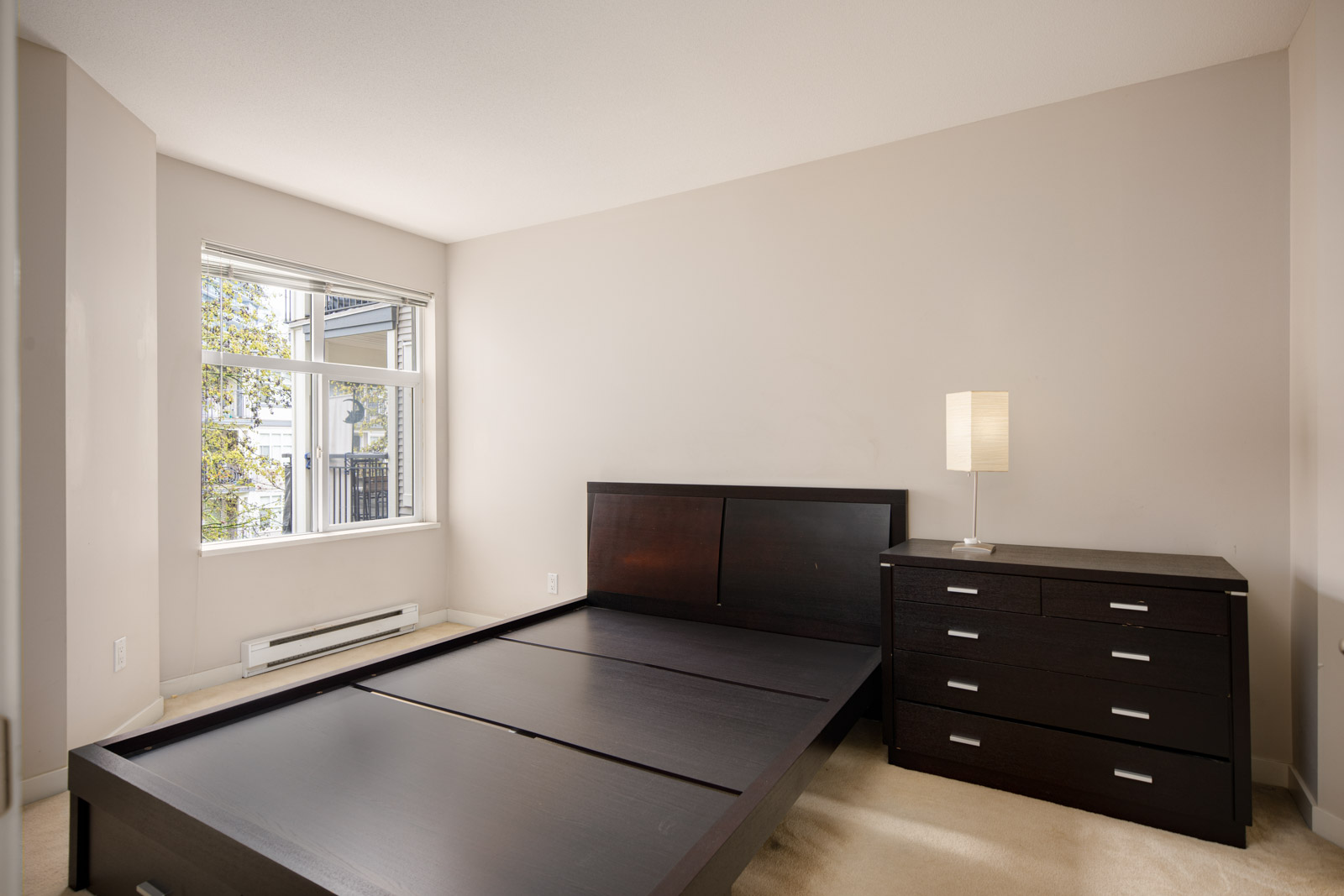 A bedroom with a large window, beige walls, an empty dark wood bed frame, a matching dresser with a lamp, and light-colored carpeted flooring.