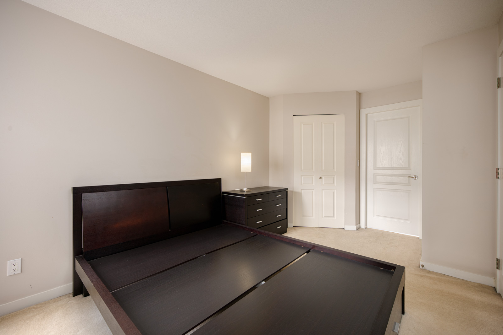 A minimally furnished bedroom with a dark wooden bedframe, matching dresser, small table lamp, beige carpet, and two white doors.