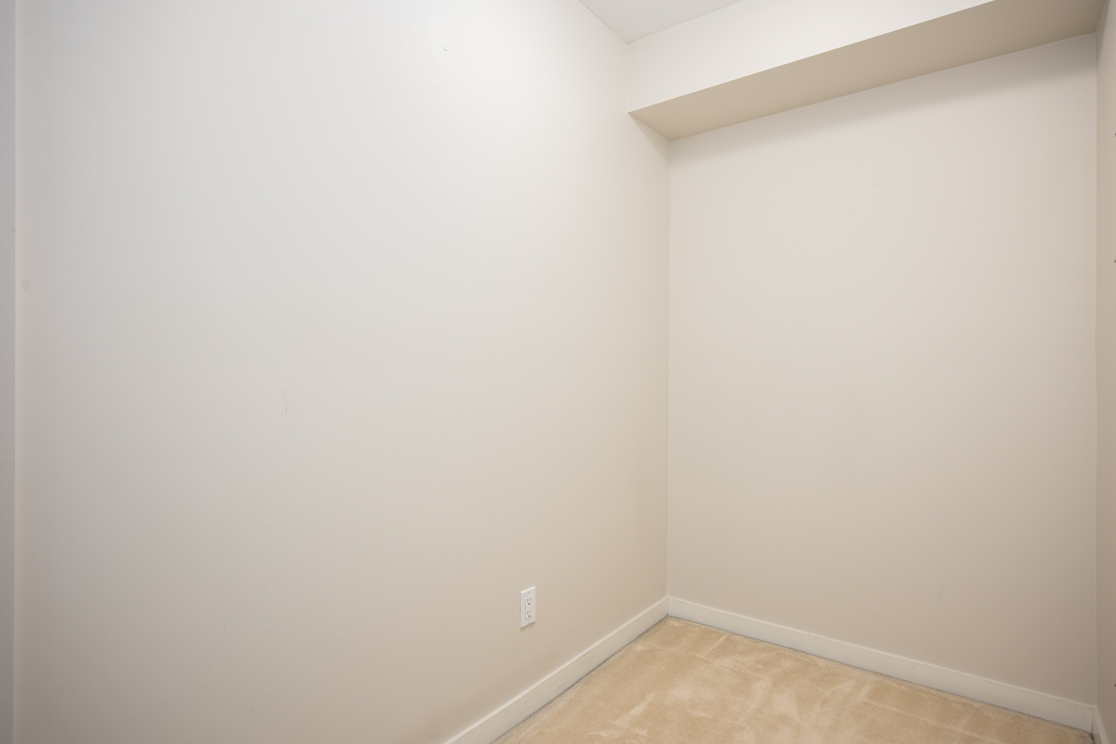 Small empty room with beige carpet, off-white walls, a single electrical outlet, and a recessed ceiling section.