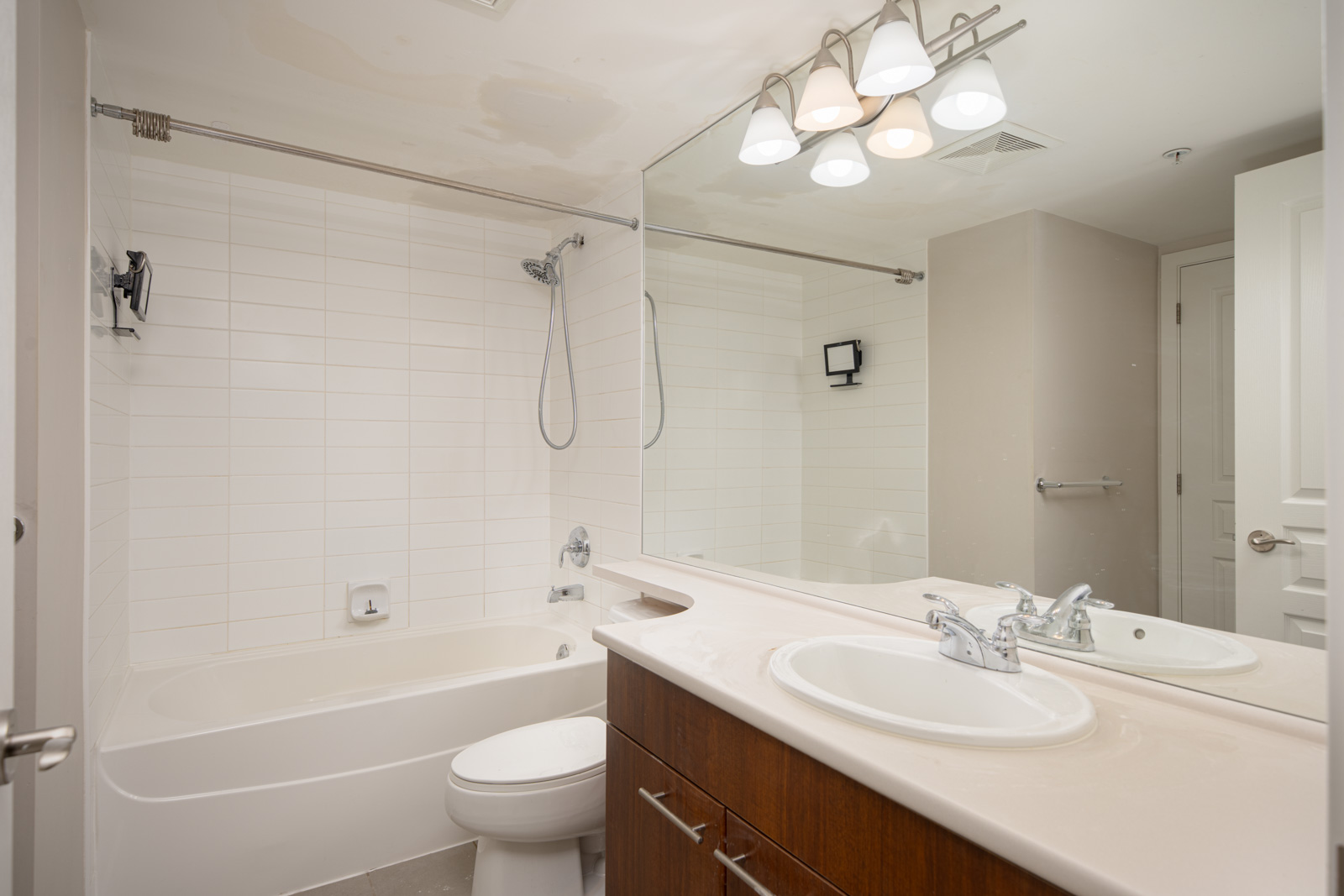 Modern bathroom with a white bathtub-shower combo, toilet, large mirror, double sinks with chrome faucets, wooden vanity, and overhead light fixture.