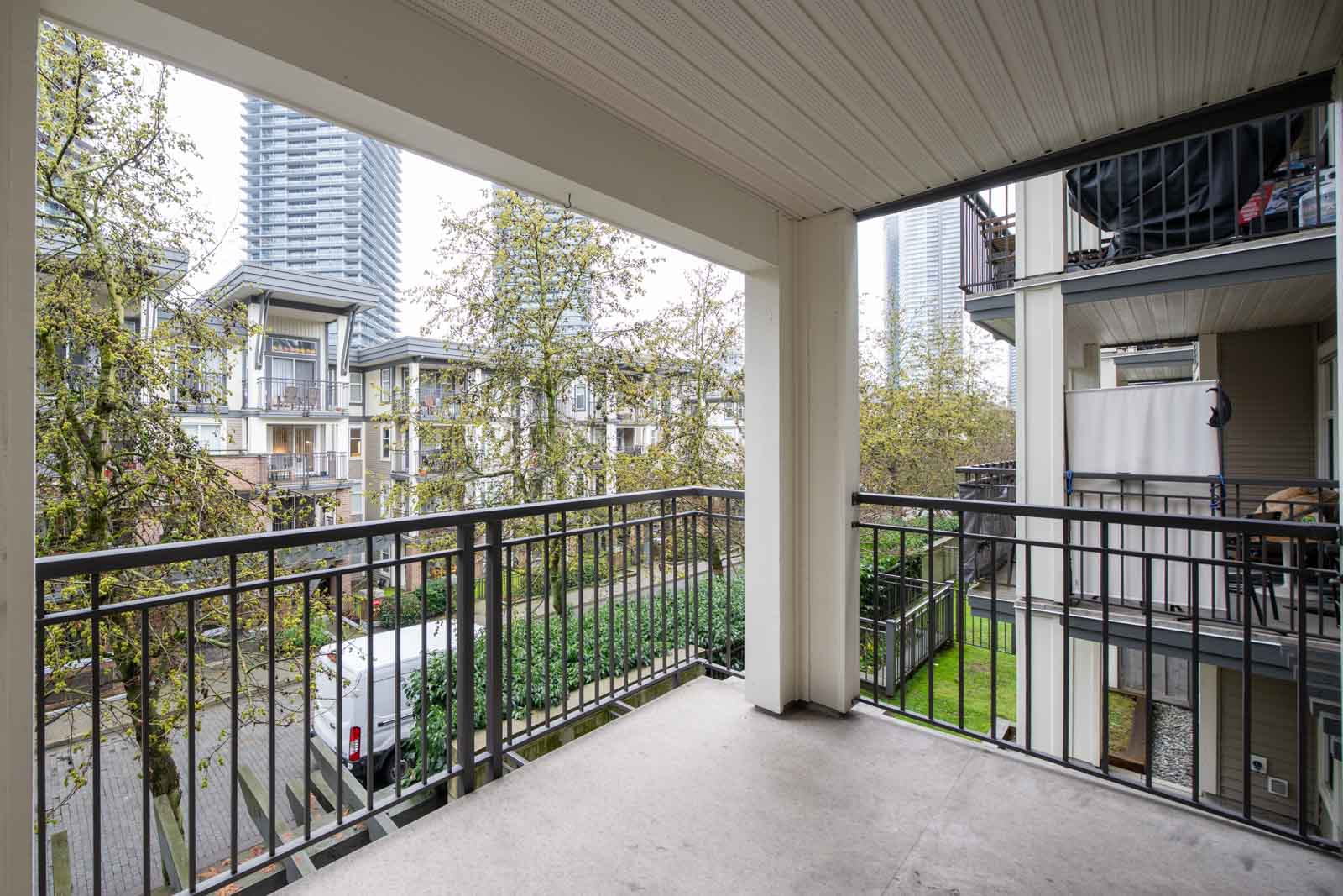 Covered apartment balcony with metal railings overlooks nearby buildings and trees in an urban setting on an overcast day.