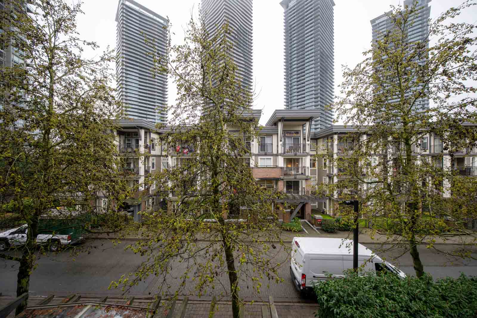 A row of mid-rise apartment buildings with trees in front, a parked white van on the street, and tall modern skyscrapers in the background.