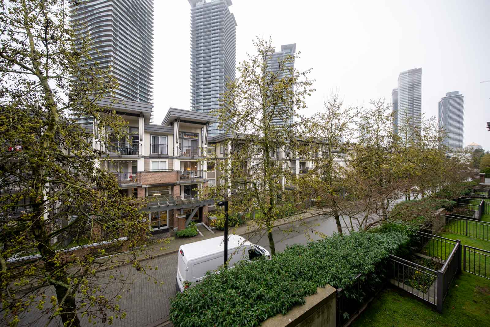 A white van is parked on a street lined with trees, with low-rise apartments and tall skyscrapers in the background on an overcast day.
