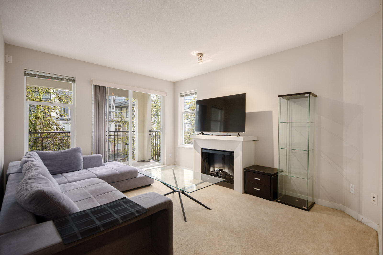 Modern living room with a gray sectional sofa, glass coffee table, TV on a white mantel, glass display cabinet, and large windows letting in natural light.