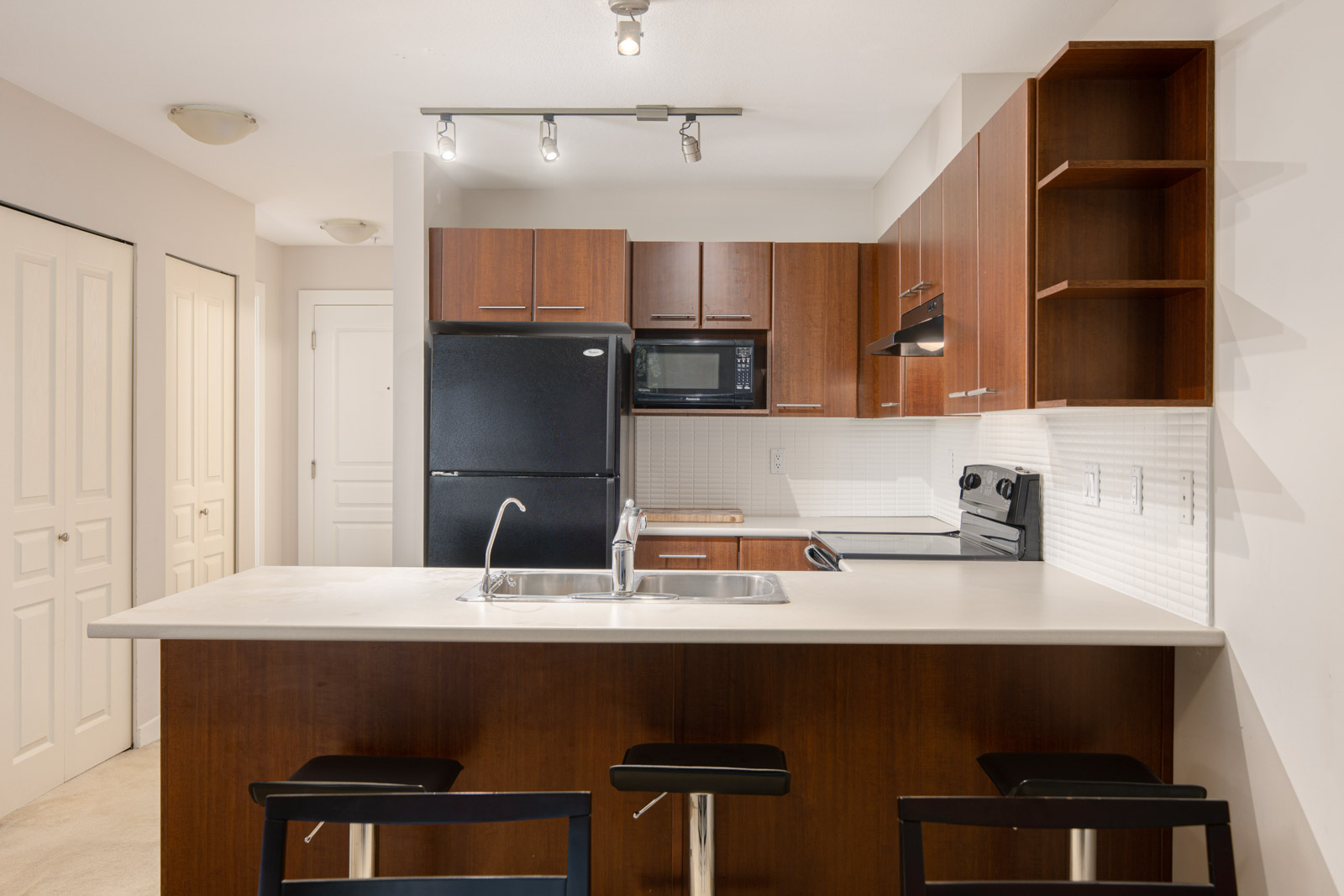 Modern kitchen with brown cabinets, black appliances, a double sink on a white countertop island, and three black bar stools.