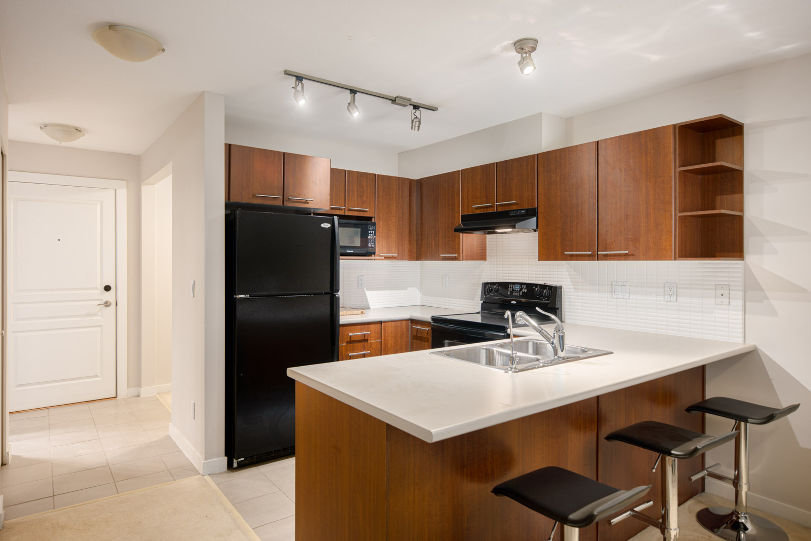 Modern kitchen with wood cabinets, black appliances, white countertops, a sink island with three bar stools, and tile flooring. The entrance door is visible in the background.