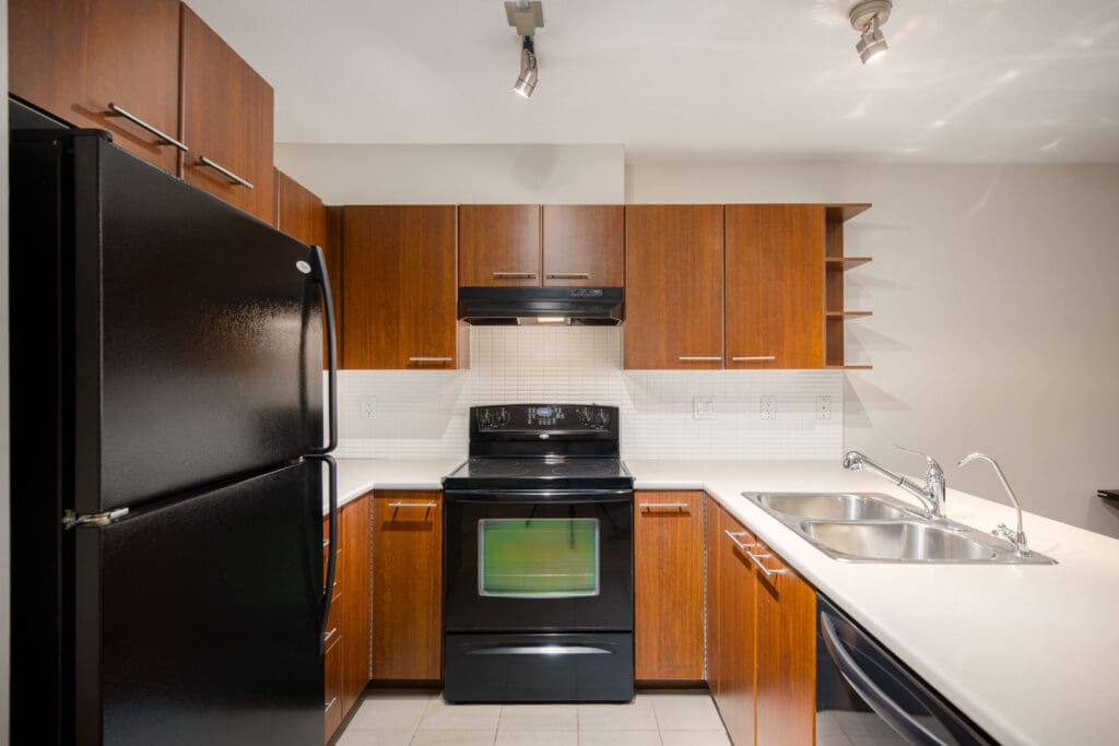 Modern kitchen with black appliances, wood cabinets, white countertops, a double sink, and tile flooring under ceiling spotlights.