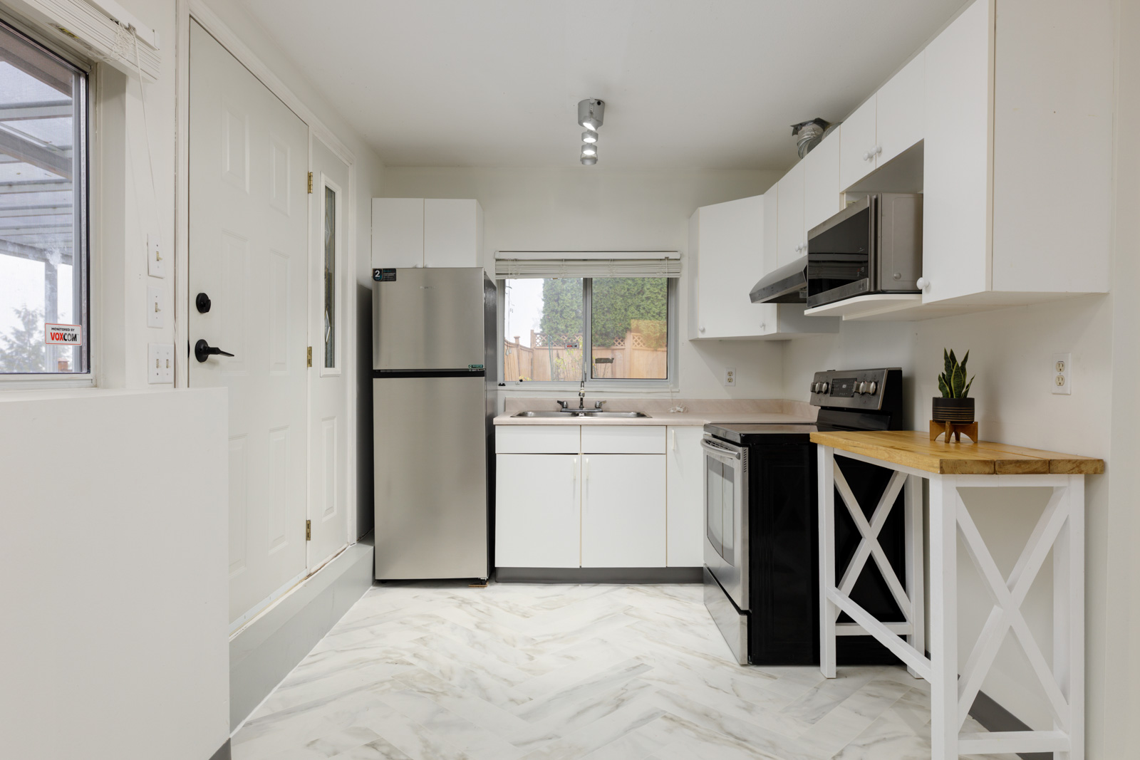 Small modern kitchen with white cabinets, stainless steel appliances, a window above the sink, and a wood-topped counter on the right. Marble-patterned floor tiles.