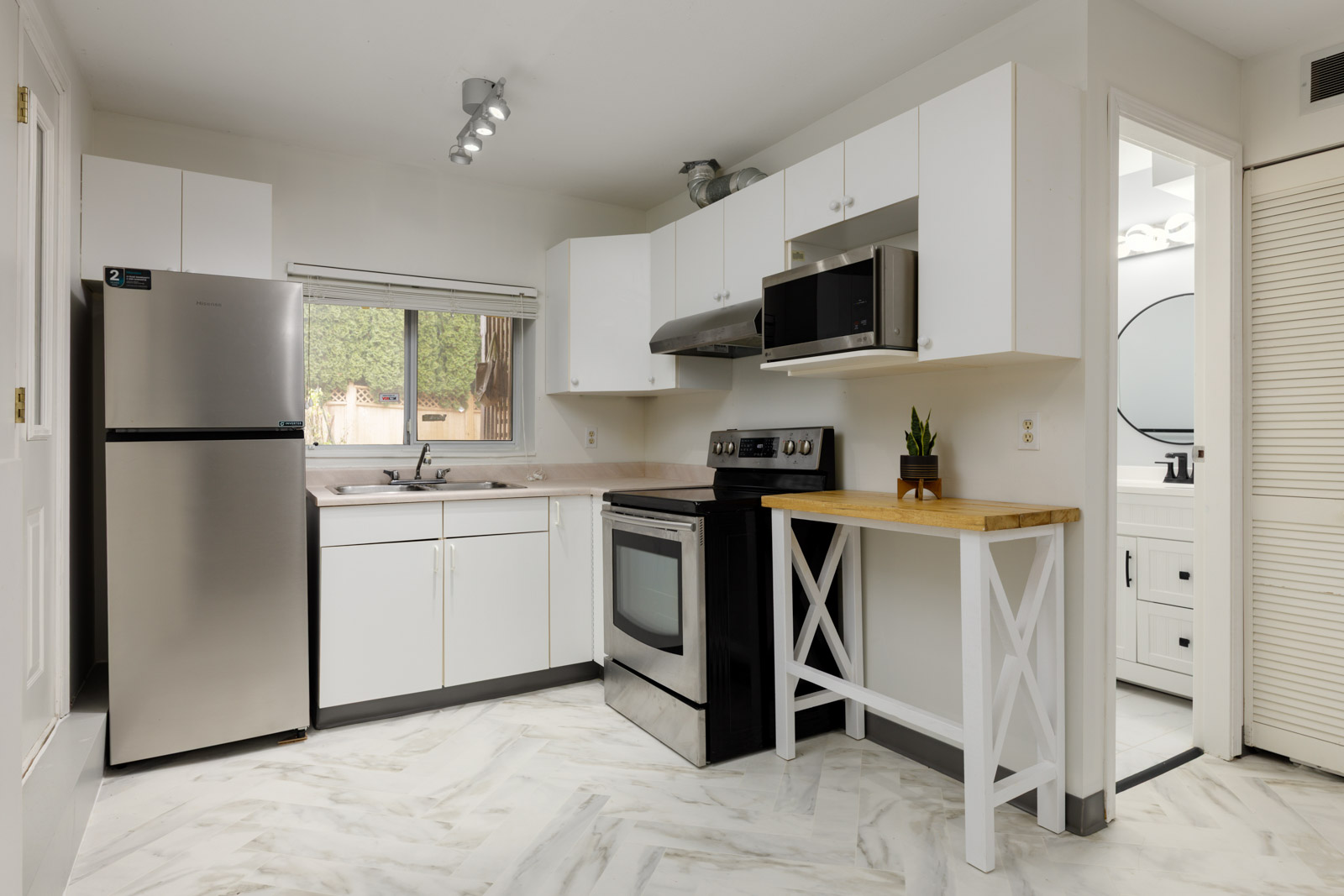 Modern kitchen with white cabinets, stainless steel refrigerator, stove, microwave, a small butcher block counter, and a window above the sink; bathroom visible in background.