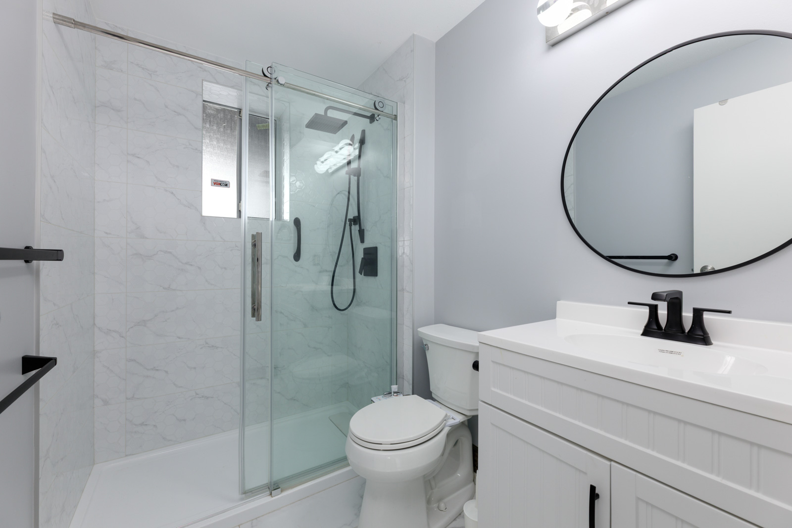 Modern bathroom with a glass-enclosed shower, white toilet, white vanity with black fixtures, and an oval mirror above the sink.