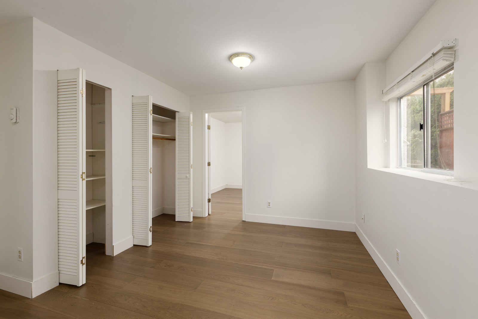 Empty room with light-colored walls, wood flooring, a ceiling light, double closet with open doors, and a window letting in natural light.