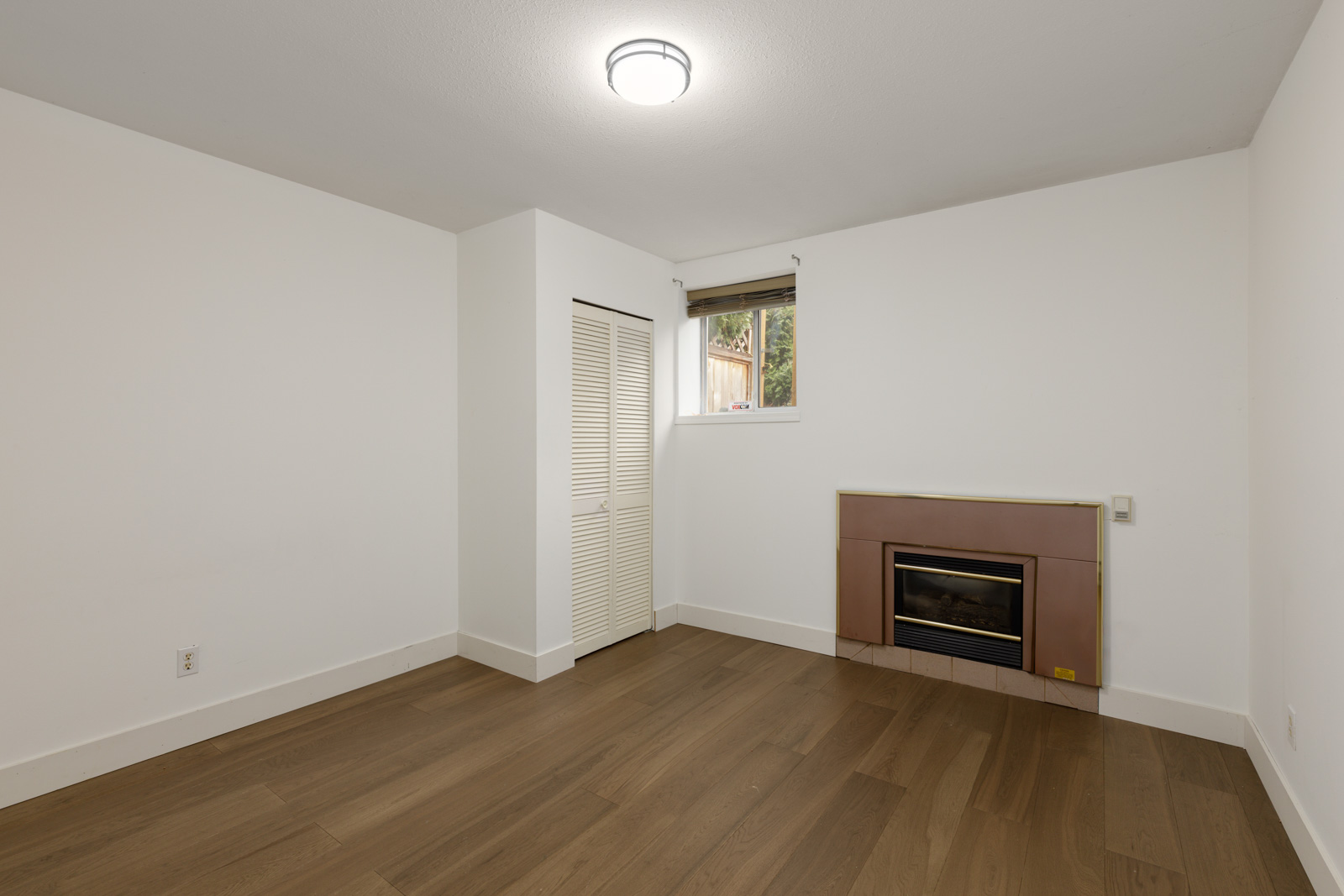 Empty room with white walls, brown wood flooring, a small window, a closed closet with louvered doors, and a fireplace with a brown surround.