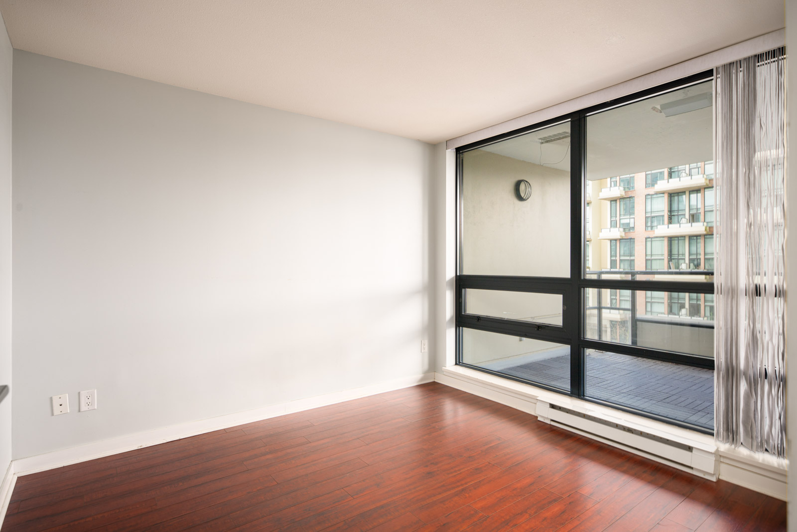 Empty room with light gray walls, dark wood floor, large window, and glass door leading to a balcony with an urban view.