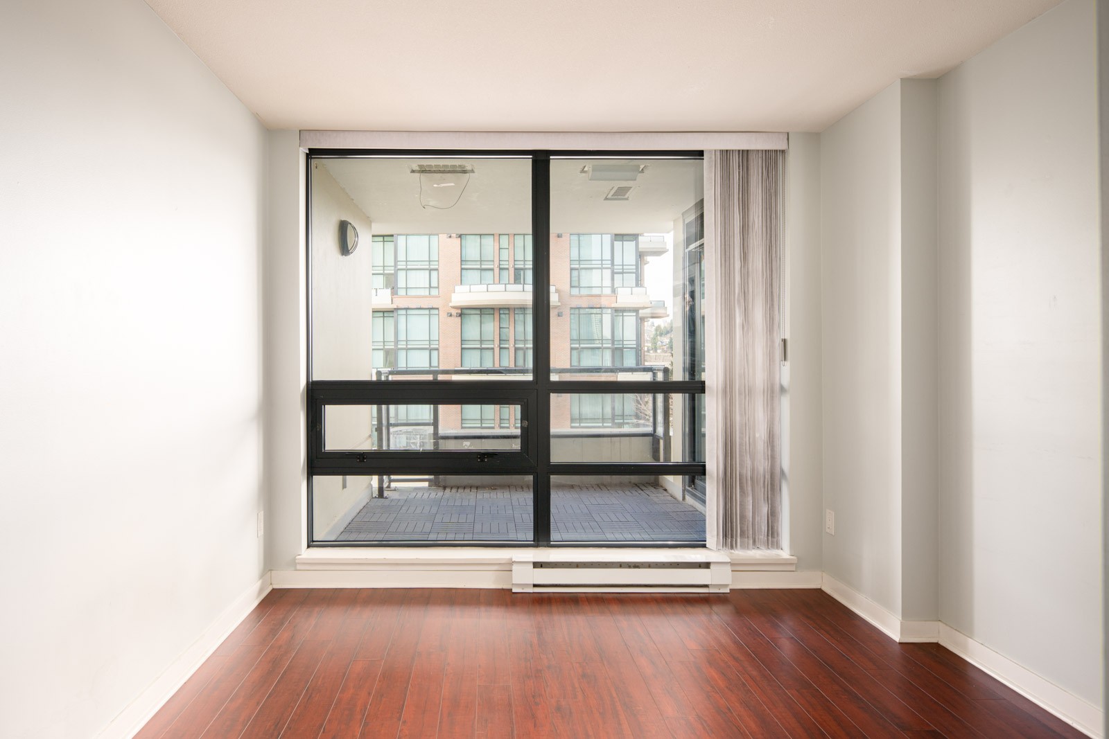 Empty room with white walls and wood flooring, featuring a large sliding glass door leading to a balcony with an apartment building visible outside.