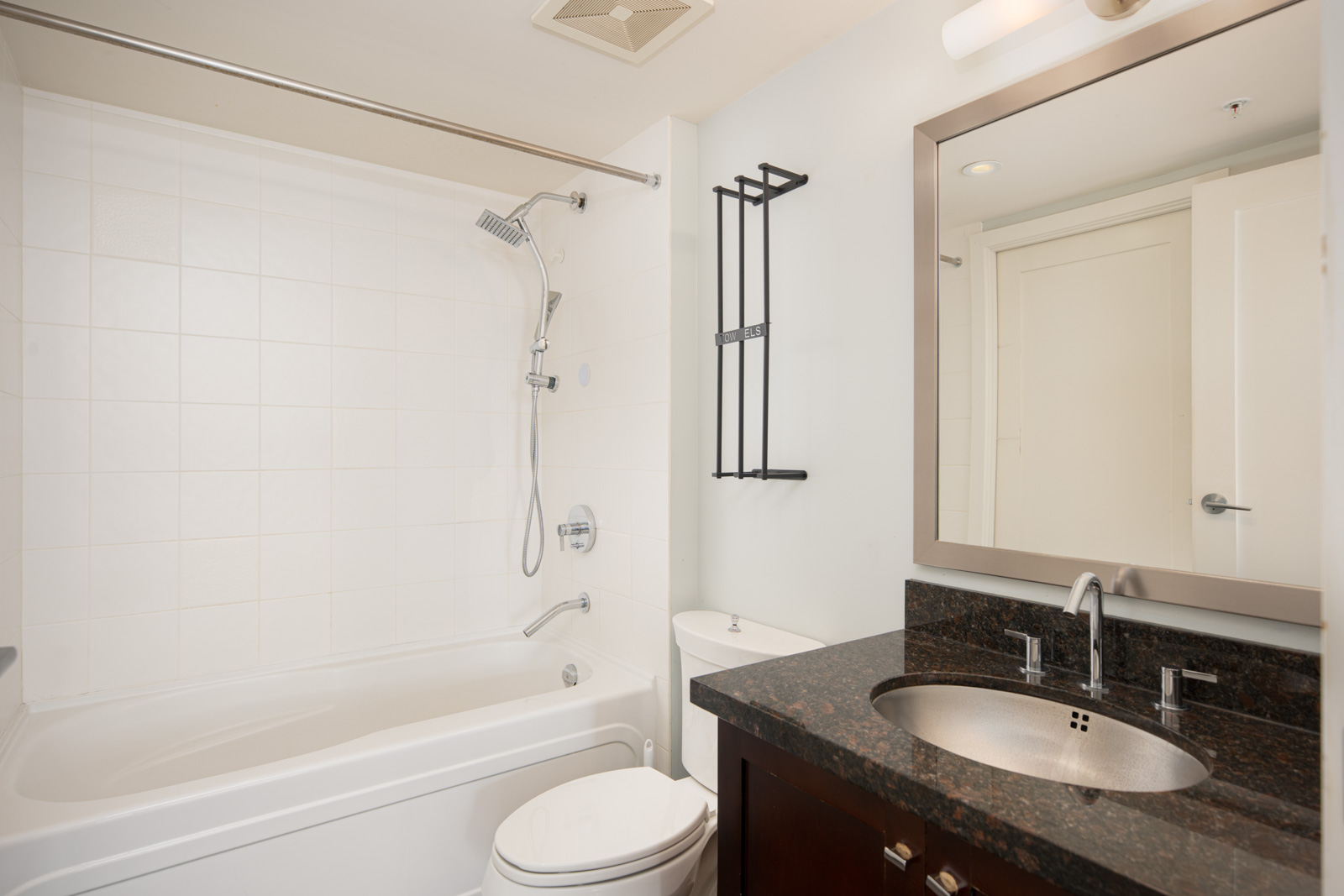Modern bathroom with a bathtub and shower, white tiled walls, a toilet, a dark countertop with a sink, a large mirror, and wall-mounted light above the mirror.