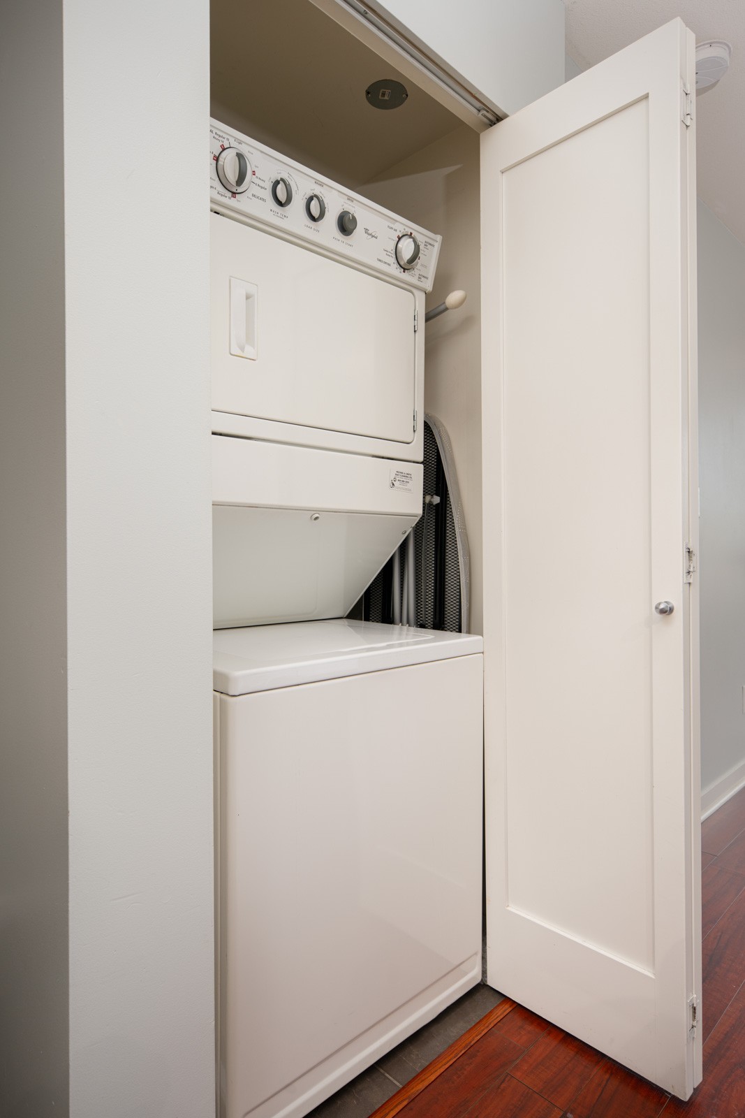 Stacked washer and dryer unit in a closet with a partially open white door, set against light-colored walls and wooden flooring.