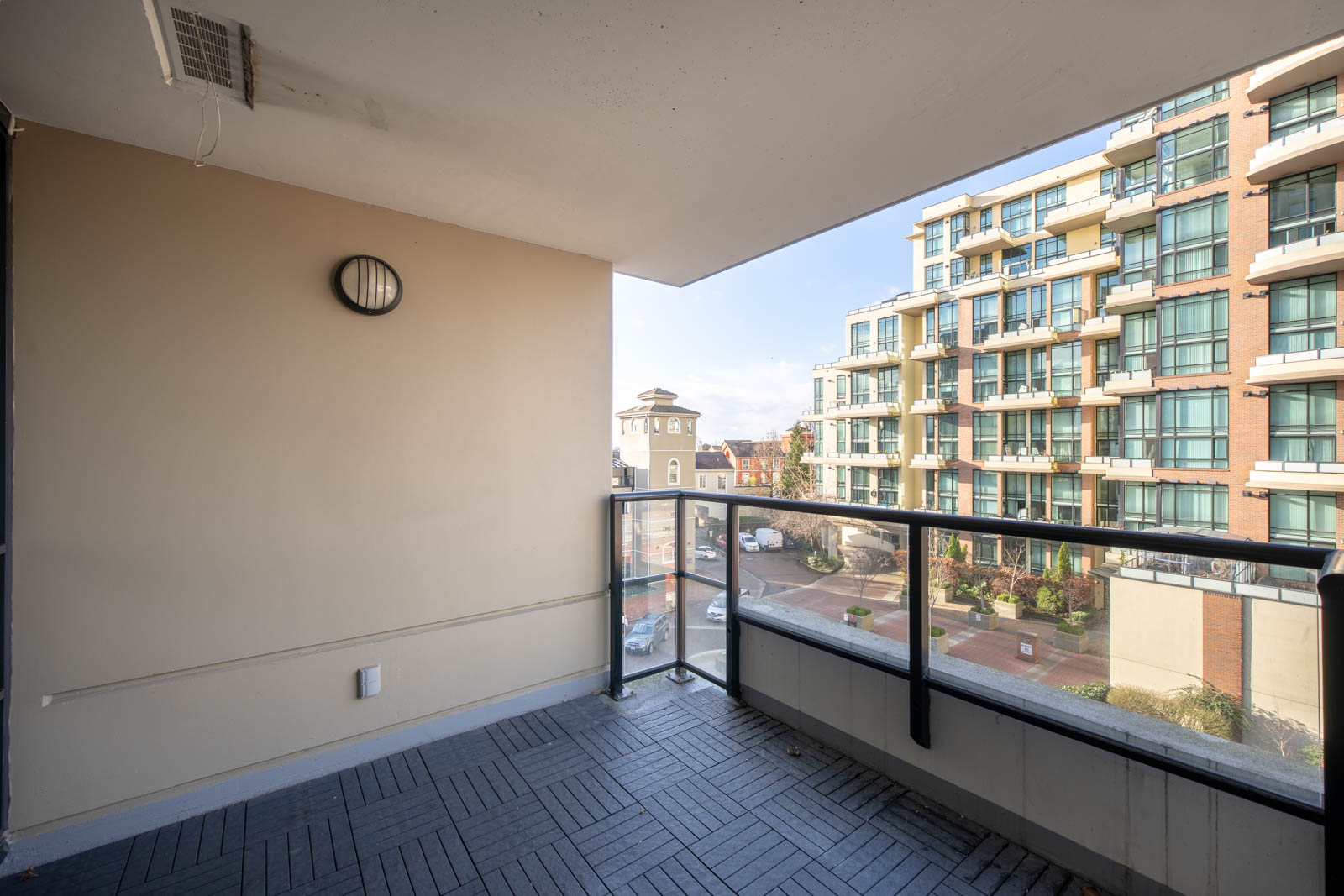 View from a modern apartment balcony with glass railing, overlooking a courtyard and neighboring residential buildings on a clear day.