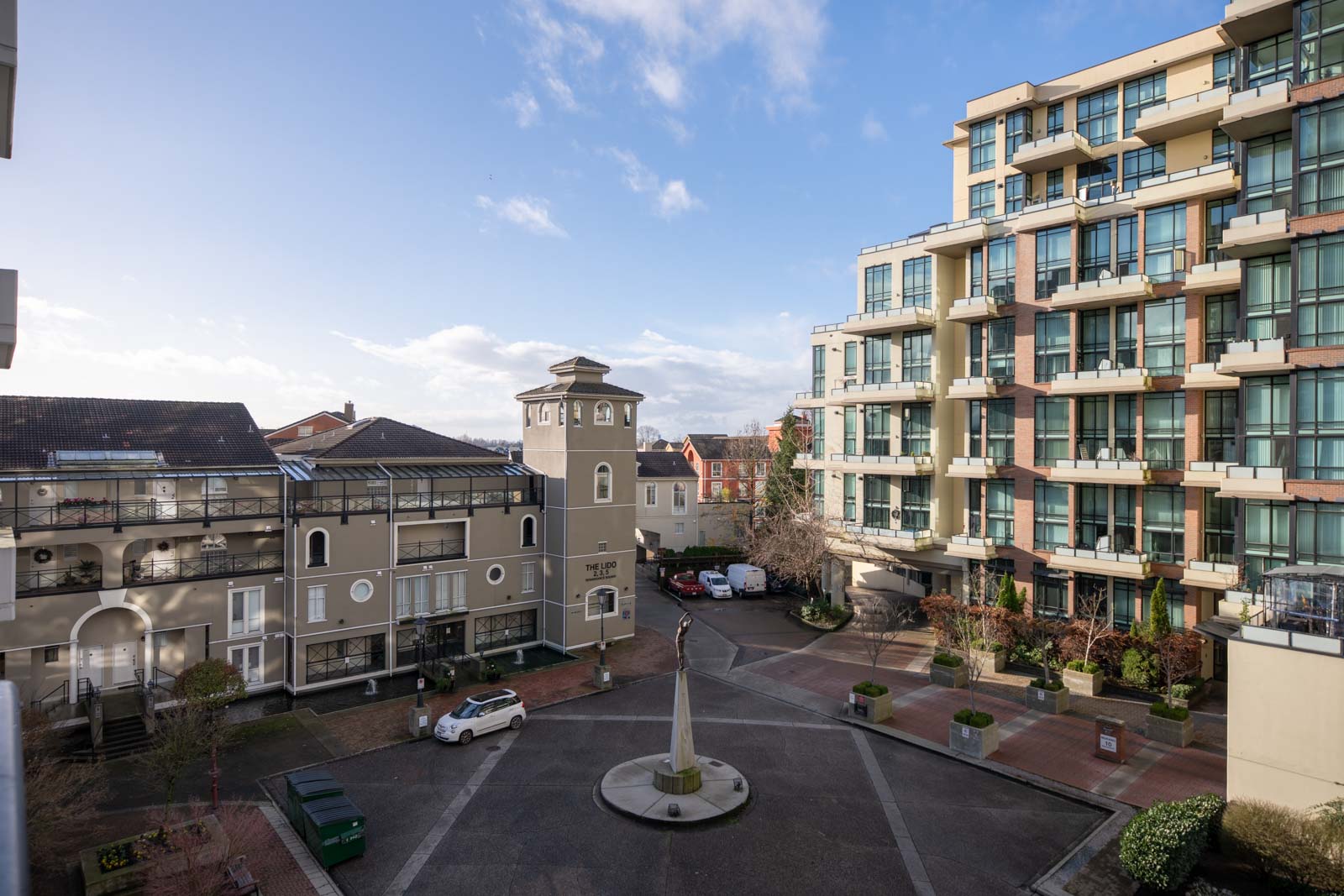 A courtyard with parked cars is surrounded by modern and historic-style apartment buildings under a clear blue sky.