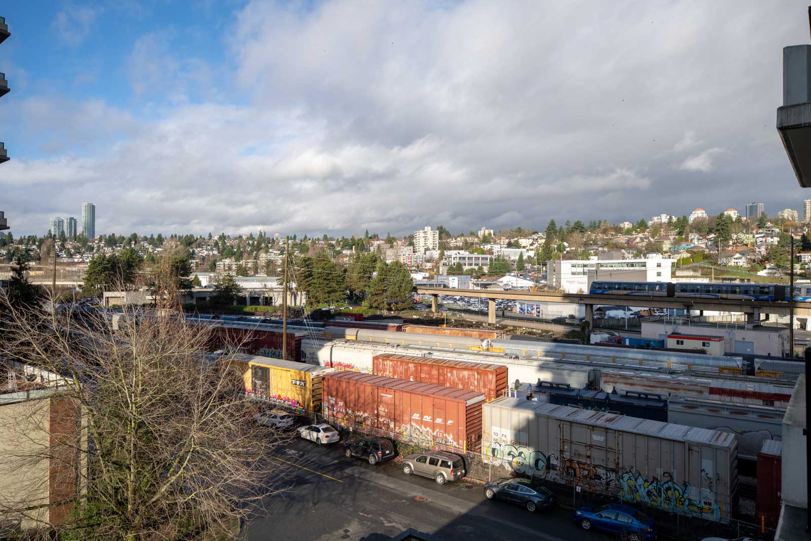 Train cars with graffiti are parked on tracks in an urban area, with a highway and city buildings in the background under a partly cloudy sky.