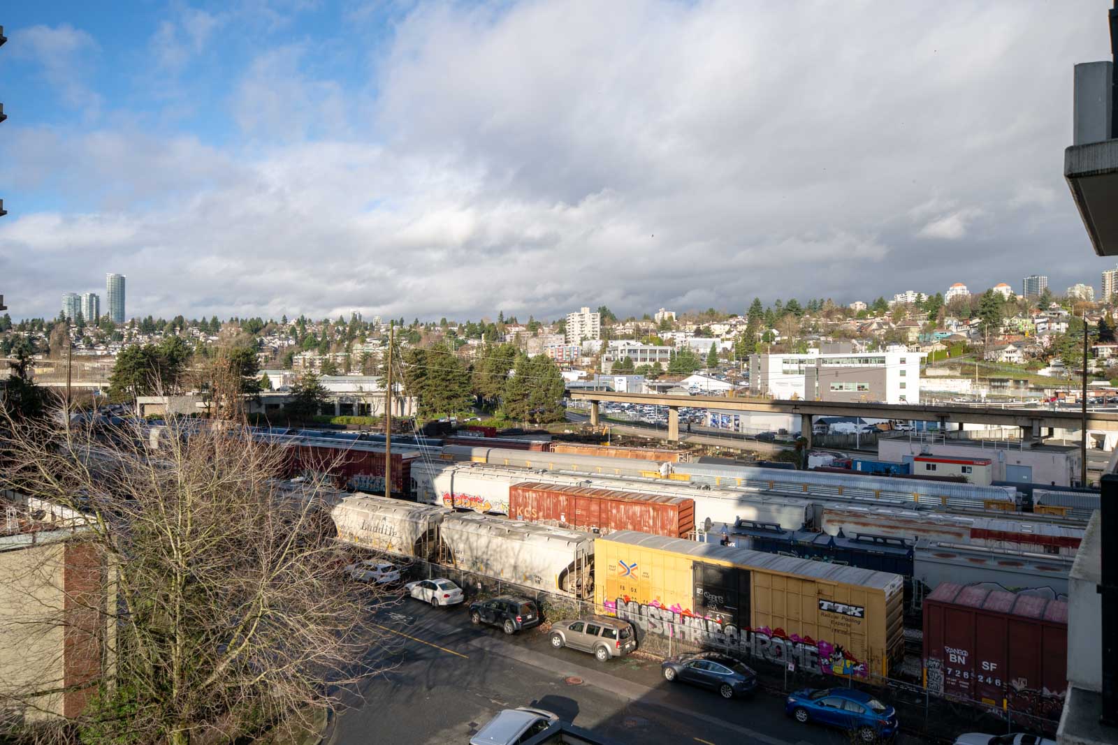 A cityscape with train cars, parked cars, and buildings under a partly cloudy sky, with distant high-rises and trees in the background.
