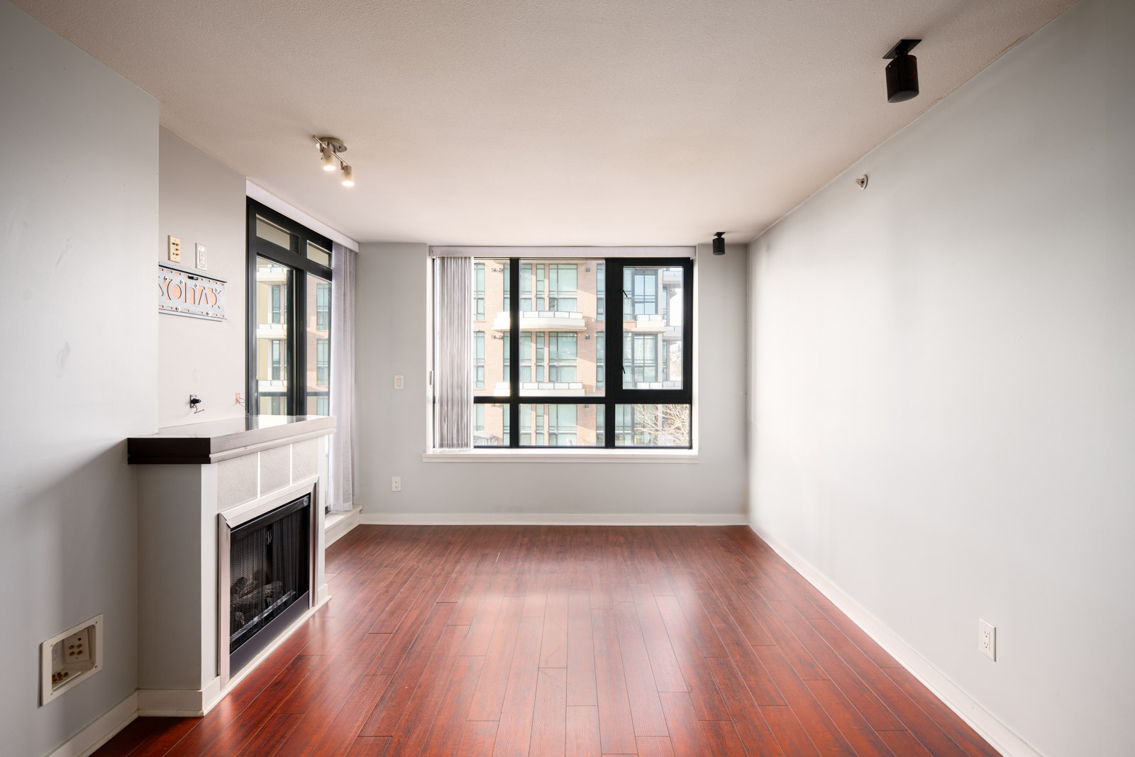 Empty room with light gray walls, dark wood floor, modern fireplace on the left, and large windows letting in natural light with a view of nearby apartment buildings.