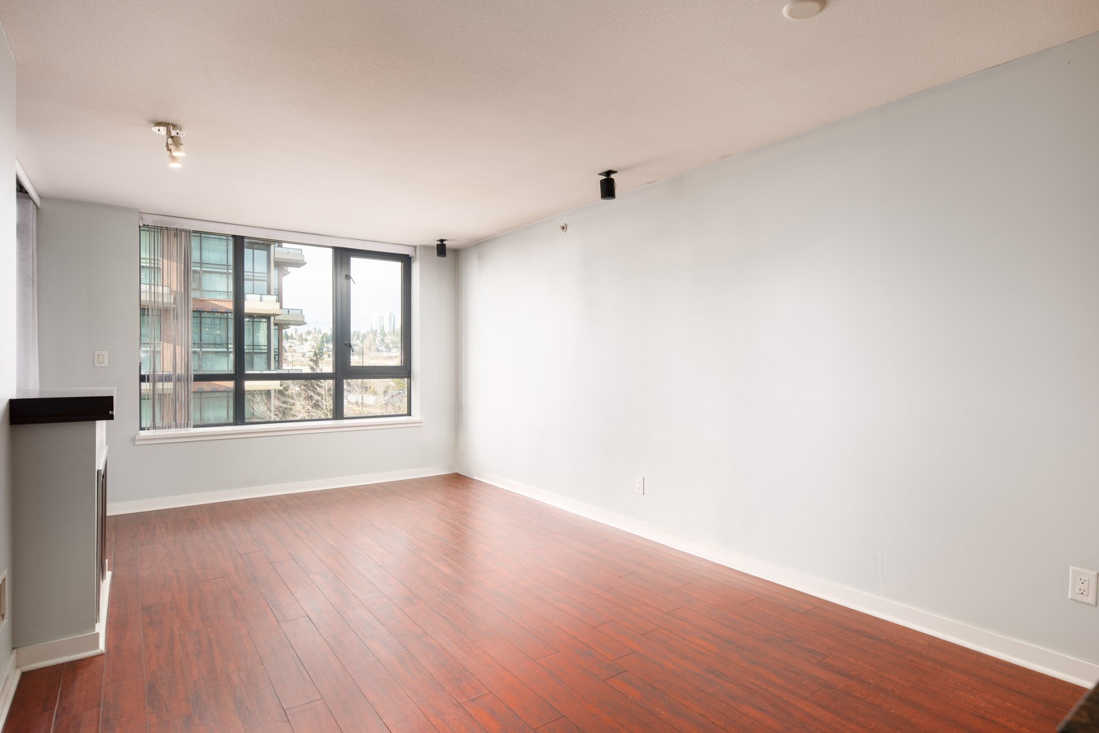 Empty living room with light gray walls, large window, and reddish-brown wood flooring. An urban building is visible through the window.