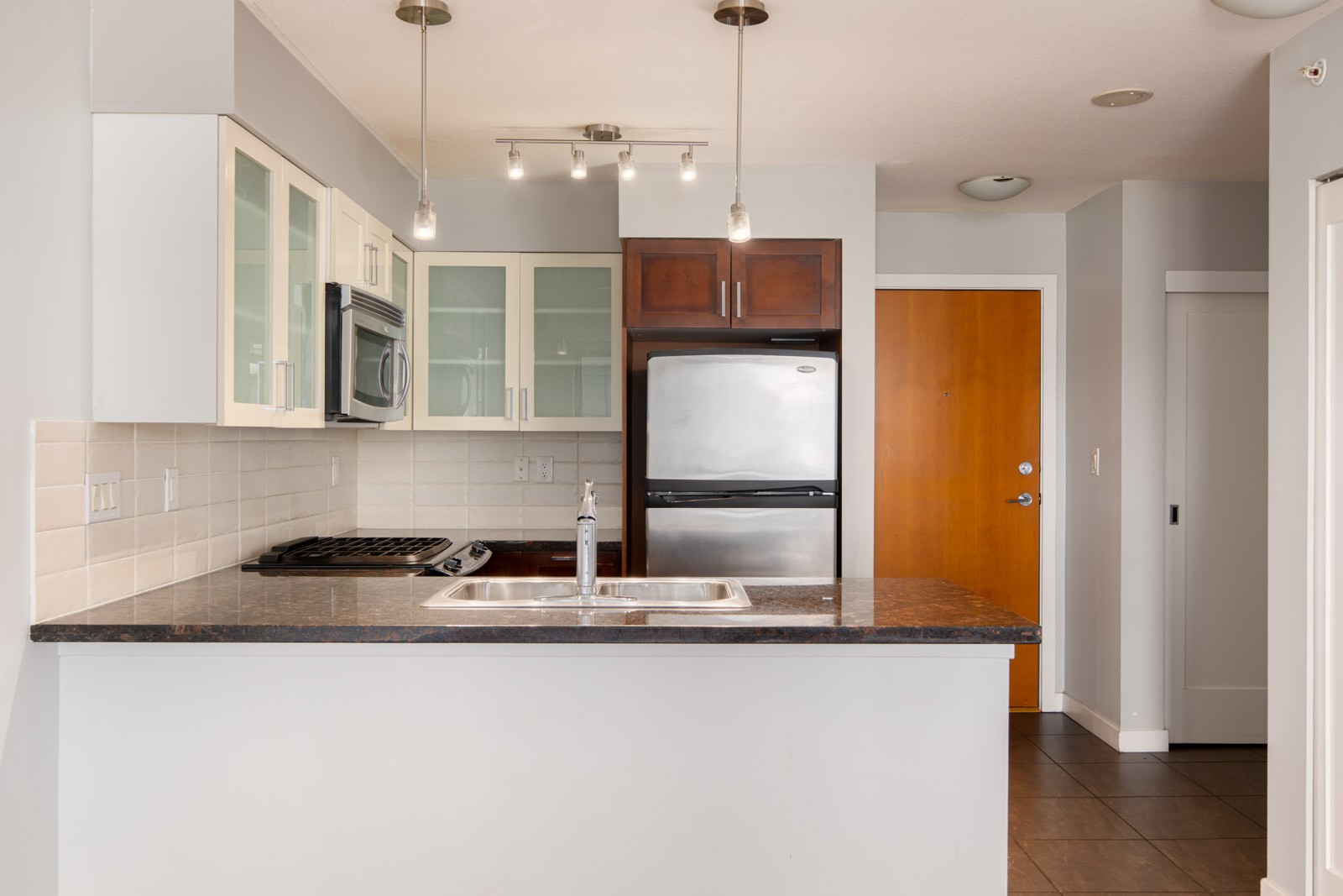 Modern kitchen with a stainless steel fridge, oven, and microwave, a double sink on a dark countertop, and light-colored cabinets against tiled backsplash.