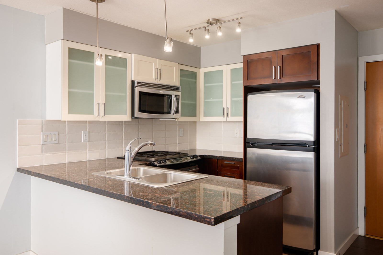 Modern kitchen with granite countertop, stainless steel appliances, dual sink, glass-front cabinets, and overhead lighting.