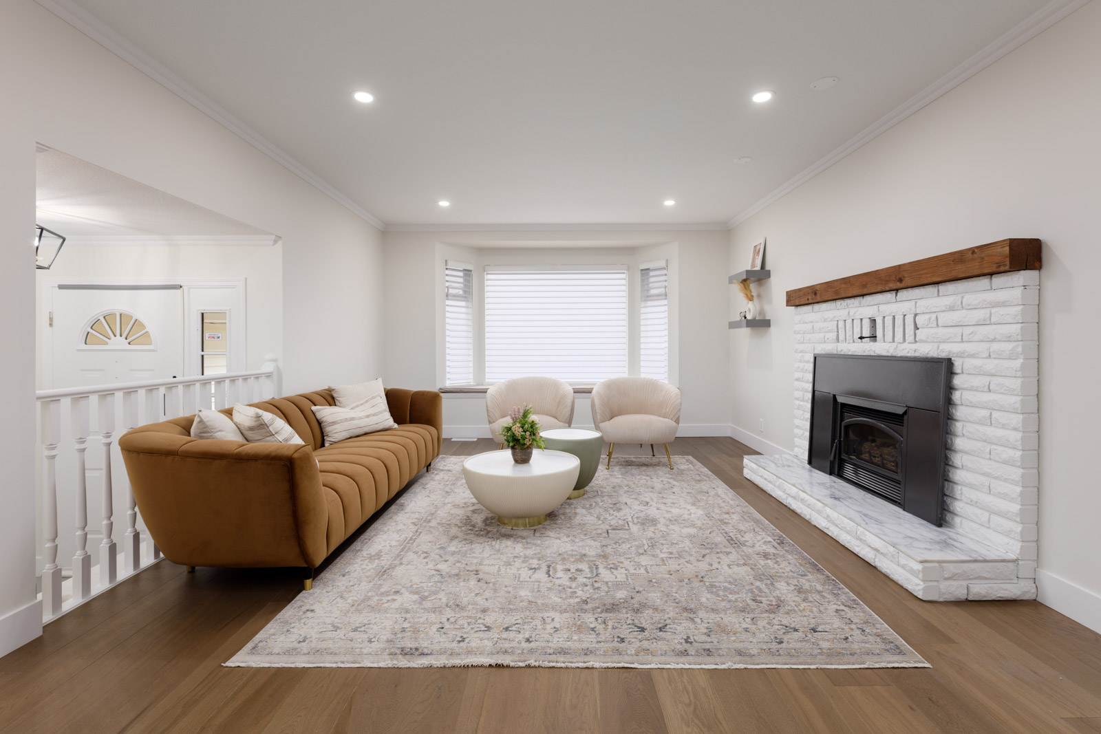 A modern living room features a brown sofa, two white chairs, a patterned rug, a white brick fireplace with a wooden mantel, and large windows with blinds.