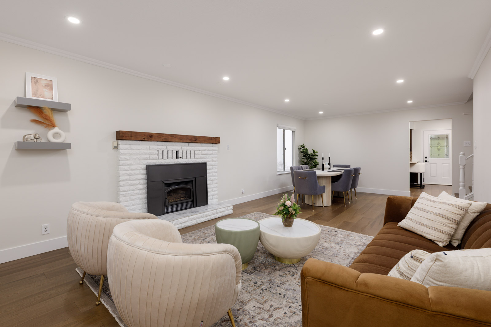 A modern living room with a brown sofa, two beige chairs, a white brick fireplace, a small dining area, and neutral décor.