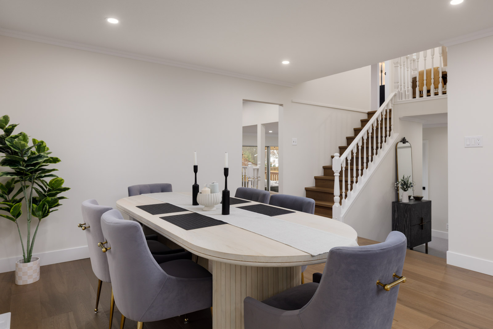Modern dining room with a light wood table, six gray chairs, table settings, a potted plant, and a staircase leading upstairs in a bright, open space.