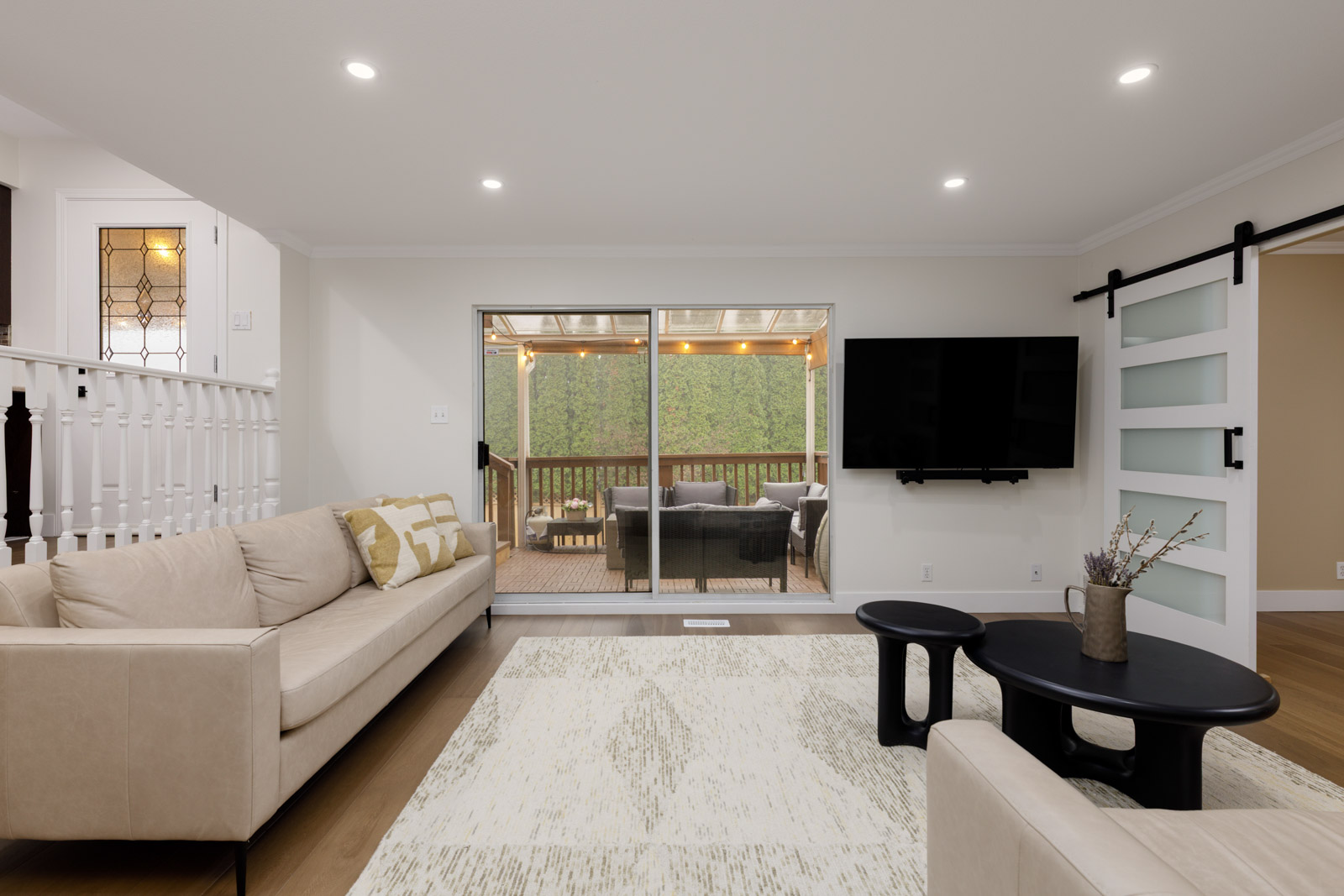 Modern living room with beige sofas, a patterned rug, black coffee tables, wall-mounted TV, and sliding glass doors leading to an outdoor patio with seating.