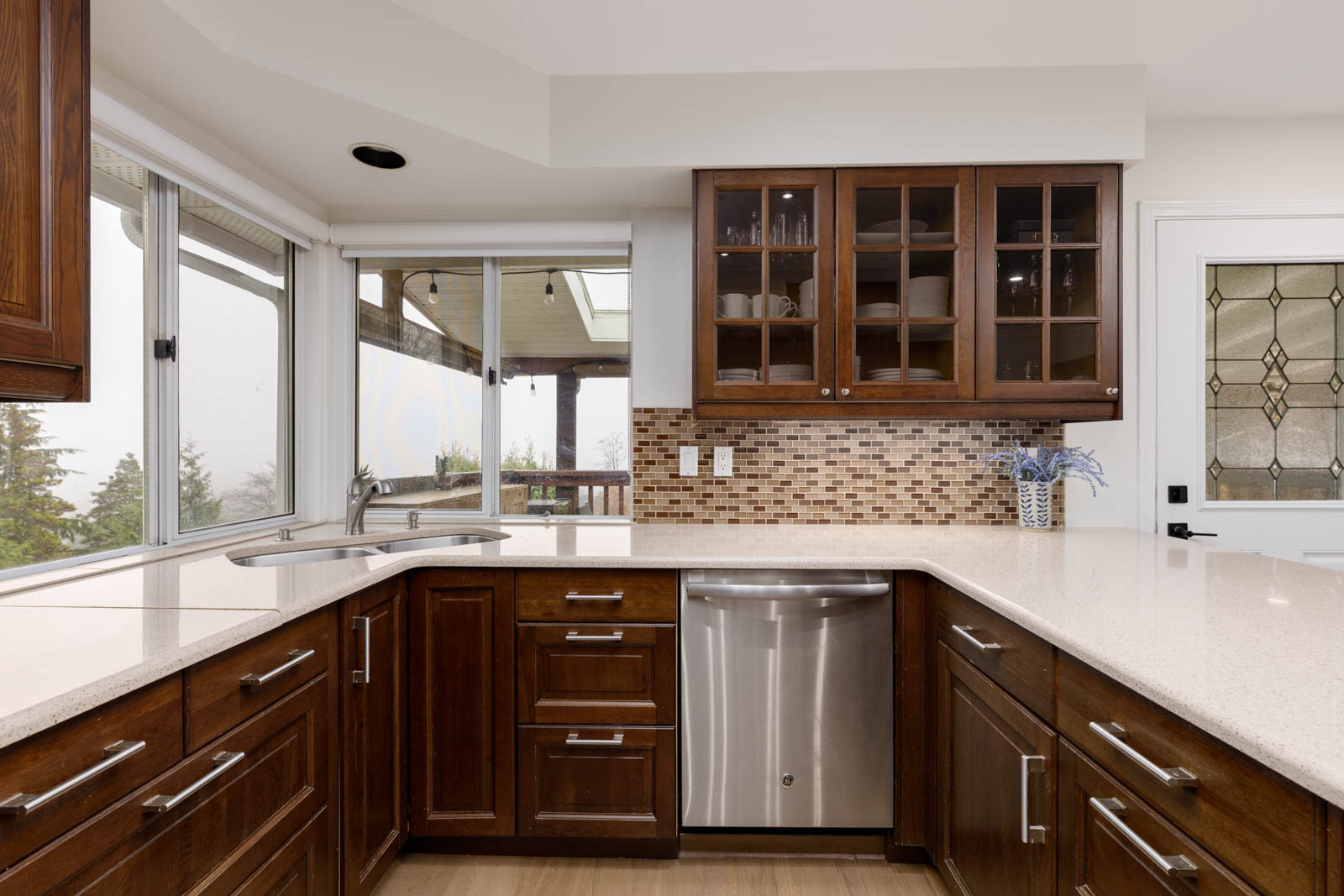 Modern kitchen with dark wood cabinets, stainless steel dishwasher, white countertops, mosaic tile backsplash, and large windows overlooking an outdoor view.