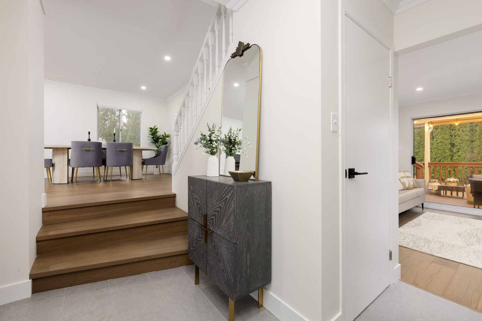 Modern entryway with a dark wood cabinet, mirror, and decorative vases; stairs lead to a dining area with purple chairs, and a living room is visible to the right.