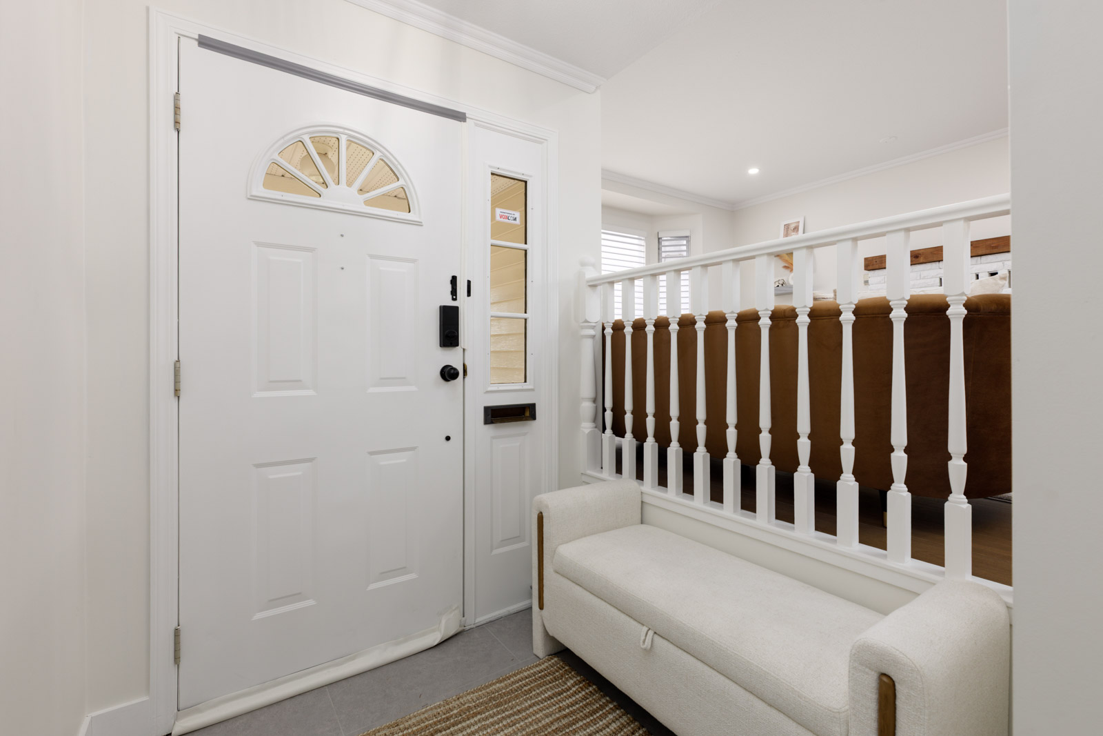 White front door with a small semi-circular window, adjacent to a bench and railing, leading to a living room area with a brown couch.