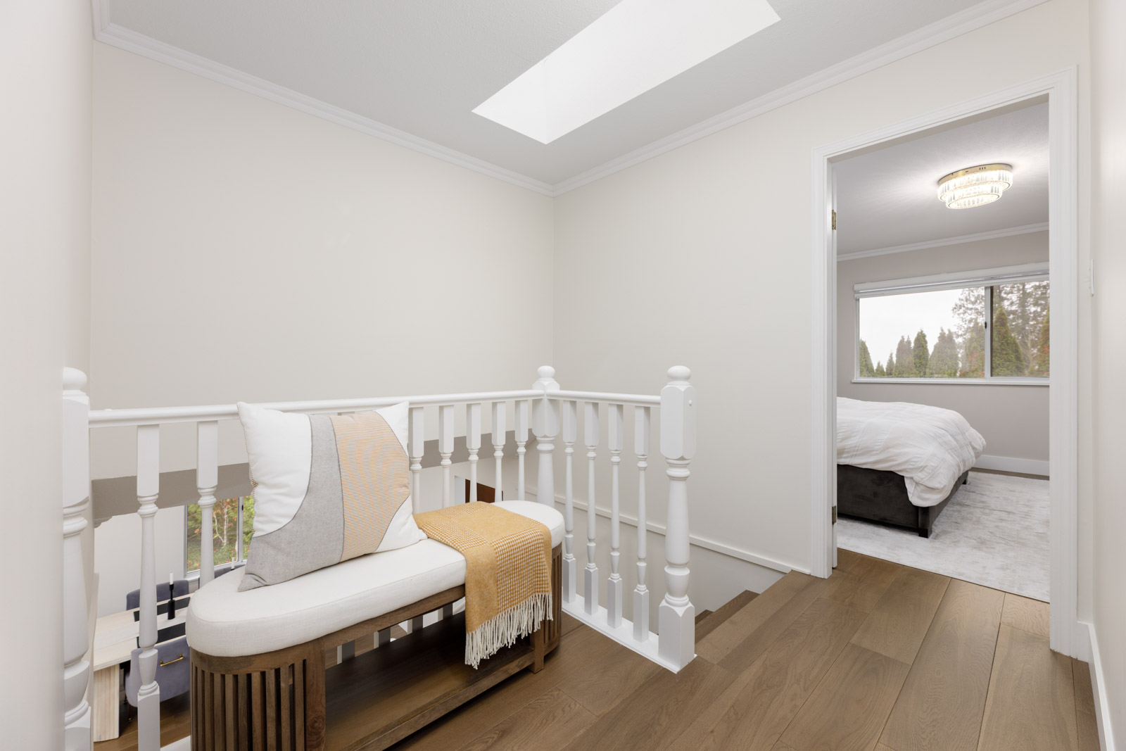 A hallway landing with a wooden bench, pillows, and a throw blanket next to a white railing, leading to a bedroom with a bed and large window.