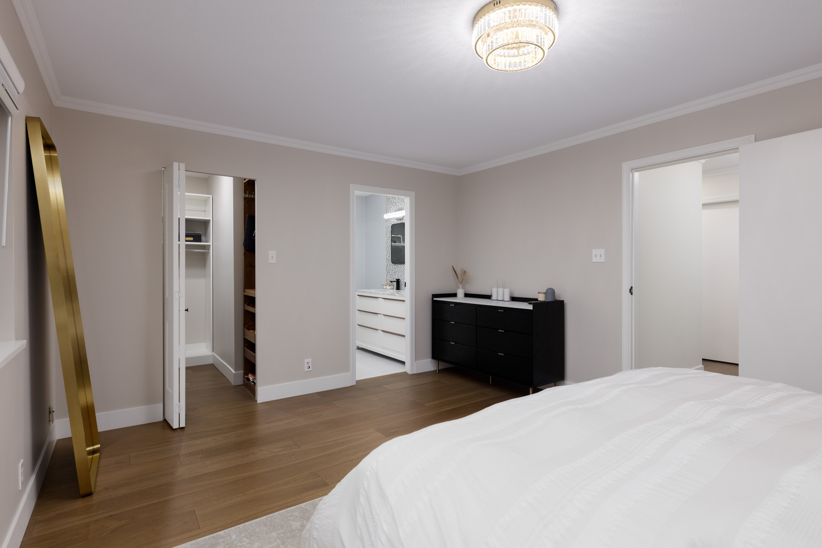 A modern bedroom with light walls, wood flooring, a bed with white bedding, a black dresser, a large mirror, and doorways to a bathroom and closet.