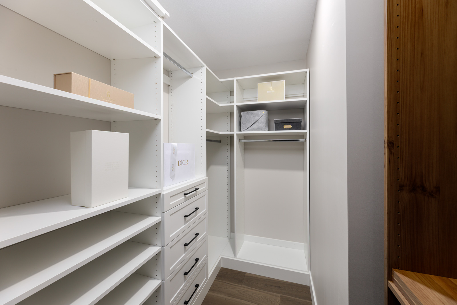 A modern walk-in closet with white shelves, drawers, and a few boxes, featuring wood flooring and empty hanging space.