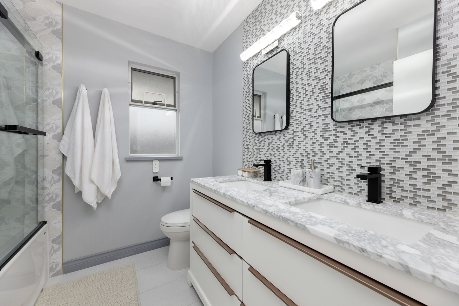 Modern bathroom with double sink vanity, marble countertop, mosaic tile backsplash, two mirrors, black faucets, toilet, towel racks, and frosted window.