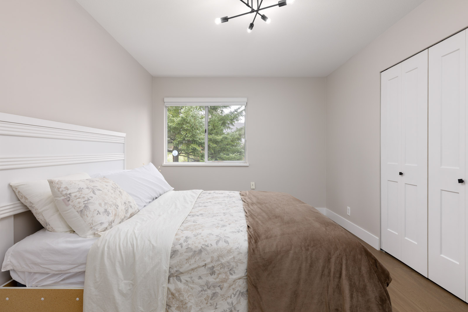 A neatly made bed with floral bedding is in a minimal bedroom with beige walls, a window, a modern ceiling light, and closed double closet doors.