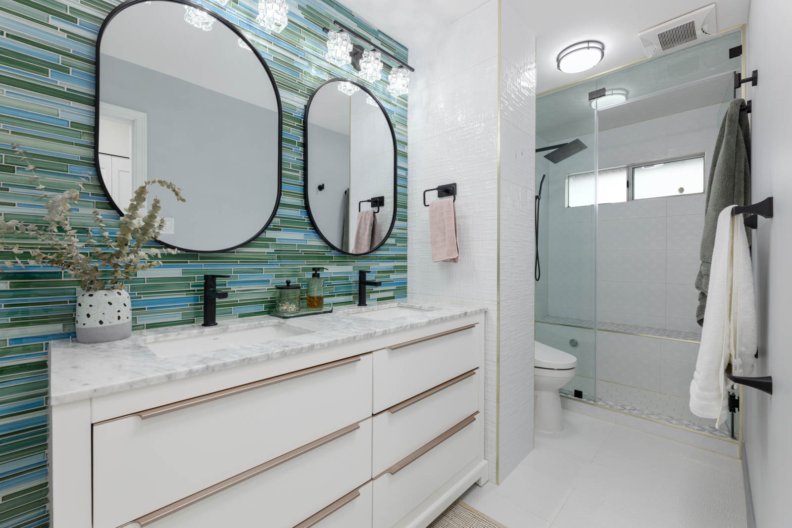 Modern bathroom with double sink vanity, oval mirrors, green and blue tiled backsplash, glass shower enclosure, and black fixtures.