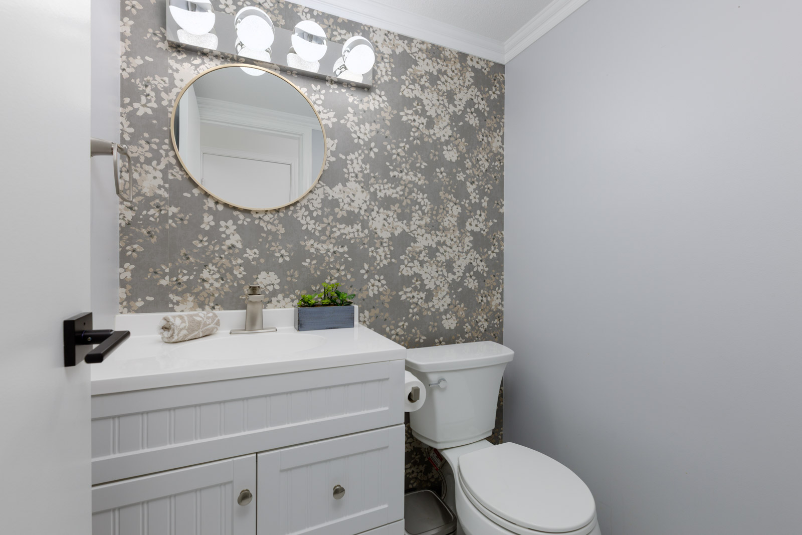 A small bathroom with a white vanity, round mirror, floral wallpaper, toilet, and overhead light fixture.