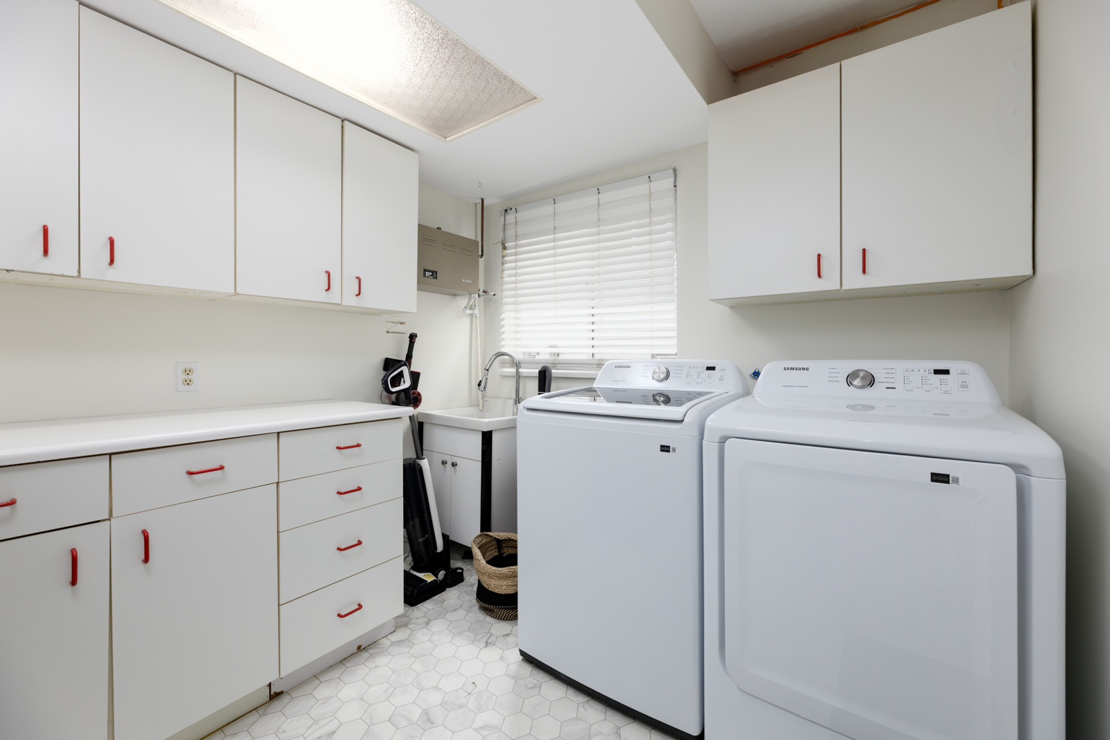A laundry room with white cabinets, a washing machine, a dryer, a utility sink, and a window with blinds.